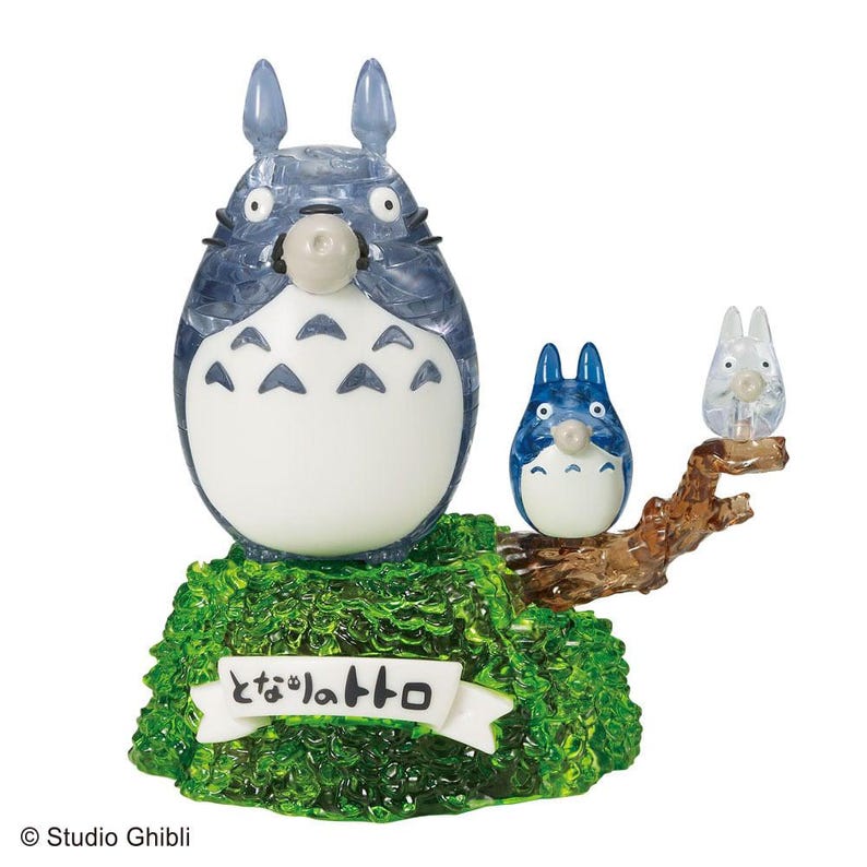 Op de afbeelding: Een doorschijnende blauw-witte Totoro-figuur met twee kleinere figuren op een bruine tak, geplaatst op een groene basis. De basis heeft een witte banner met Japanse tekst. De figuren zijn van de Studio Ghibli-collectie.