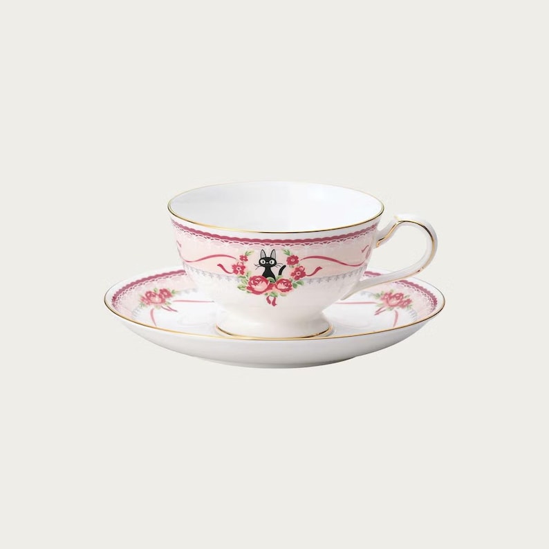 May include: A white teacup and saucer set with a pink floral design and gold trim. The teacup features a black cat illustration surrounded by roses. The saucer has a matching floral pattern, creating a charming and elegant tea set.