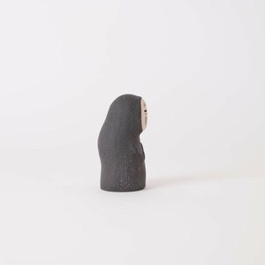 Puede incluir: Una peque&ntilde;a figura de cer&aacute;mica gris oscuro con textura moteada. La figura tiene una forma redondeada y encapuchada con un rostro p&aacute;lido y liso que presenta dos ojos negros simples y una peque&ntilde;a boca negra, vista de perfil sobre fondo liso.