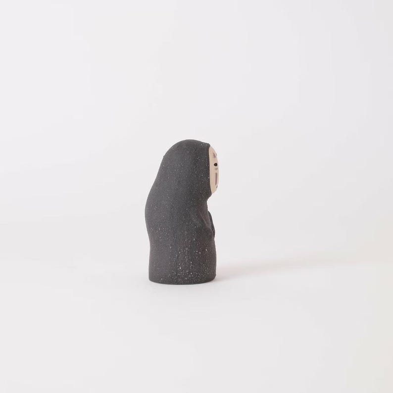 Puede incluir: Una peque&ntilde;a figura de cer&aacute;mica gris oscuro con textura moteada. La figura tiene una forma redondeada y encapuchada con un rostro p&aacute;lido y liso que presenta dos ojos negros simples y una peque&ntilde;a boca negra, vista de perfil sobre fondo liso.