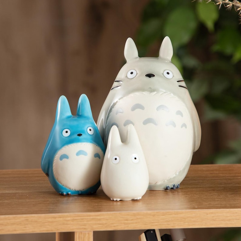 May include: Three ceramic Totoro figurines in varying sizes. The largest is gray with black markings, the medium is blue, and the smallest is white. All have large eyes and are standing on a wooden surface.