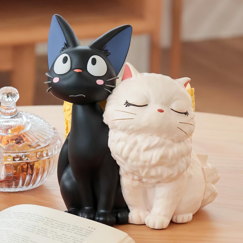 Puede incluir: Una figura decorativa con dos gatos sentados uno al lado del otro. Un gato es negro con ojos blancos grandes, orejas interiores azules y manchas rosas en las mejillas. El otro gato es blanco de pelo largo, ojos cerrados y nariz peque&ntilde;a rosa.