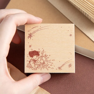 May include: A square wooden rubber stamp featuring a dark brown engraved illustration of a person with short hair holding a glowing orb, surrounded by scattered starbursts and two shooting stars across the top and bottom corners of the surface.