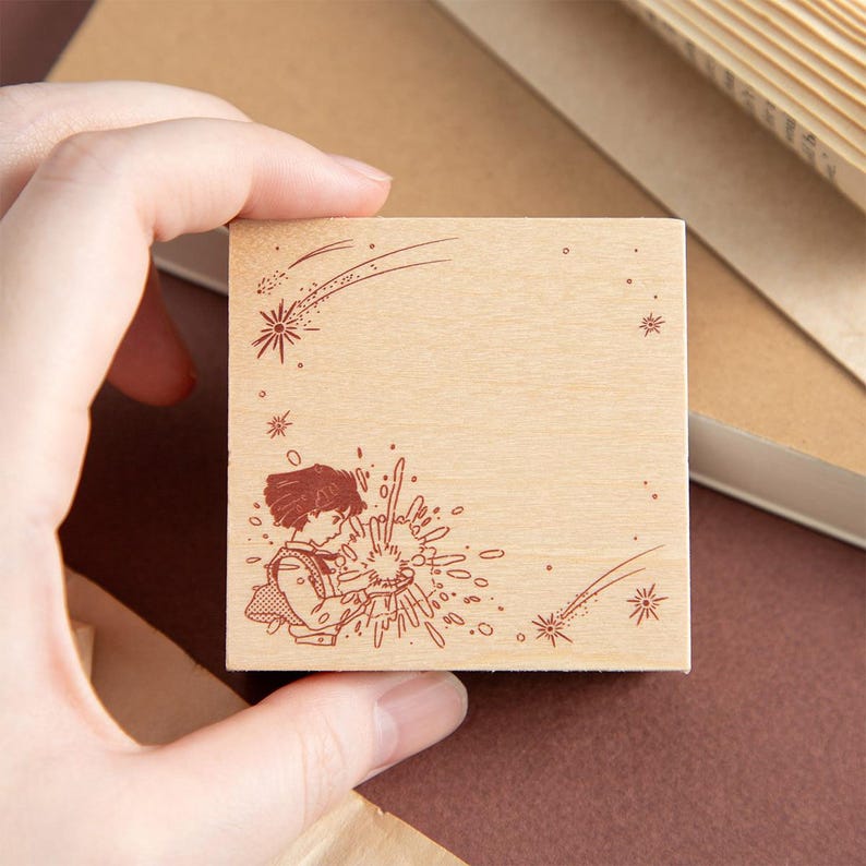 May include: A square wooden rubber stamp featuring a dark brown engraved illustration of a person with short hair holding a glowing orb, surrounded by scattered starbursts and two shooting stars across the top and bottom corners of the surface.