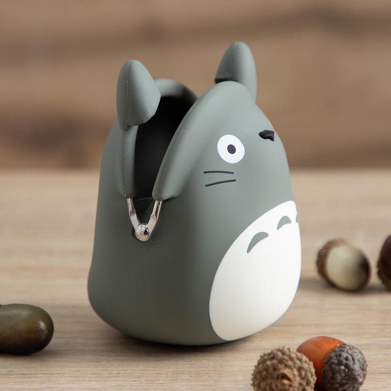 May include: A gray coin purse shaped like a cartoon character, with a white belly and black details. The purse has a silver clasp and two small ears. The background includes acorns on a wooden surface.
