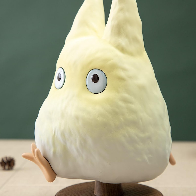 May include: A light yellow, cartoon-like figurine with large, round eyes and small, outstretched arms. The figurine has a fluffy texture and is mounted on a small, round, wooden base. The background is a solid, muted green.