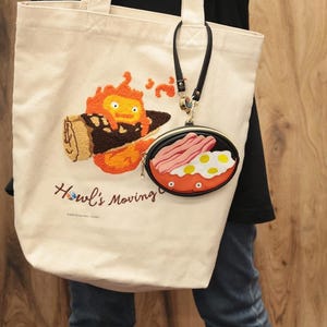 May include: Cream tote bag featuring an embroidered orange fiery character and the text "Howl's Moving". A small, round coin purse with a fried egg and bacon design is attached to the bag.