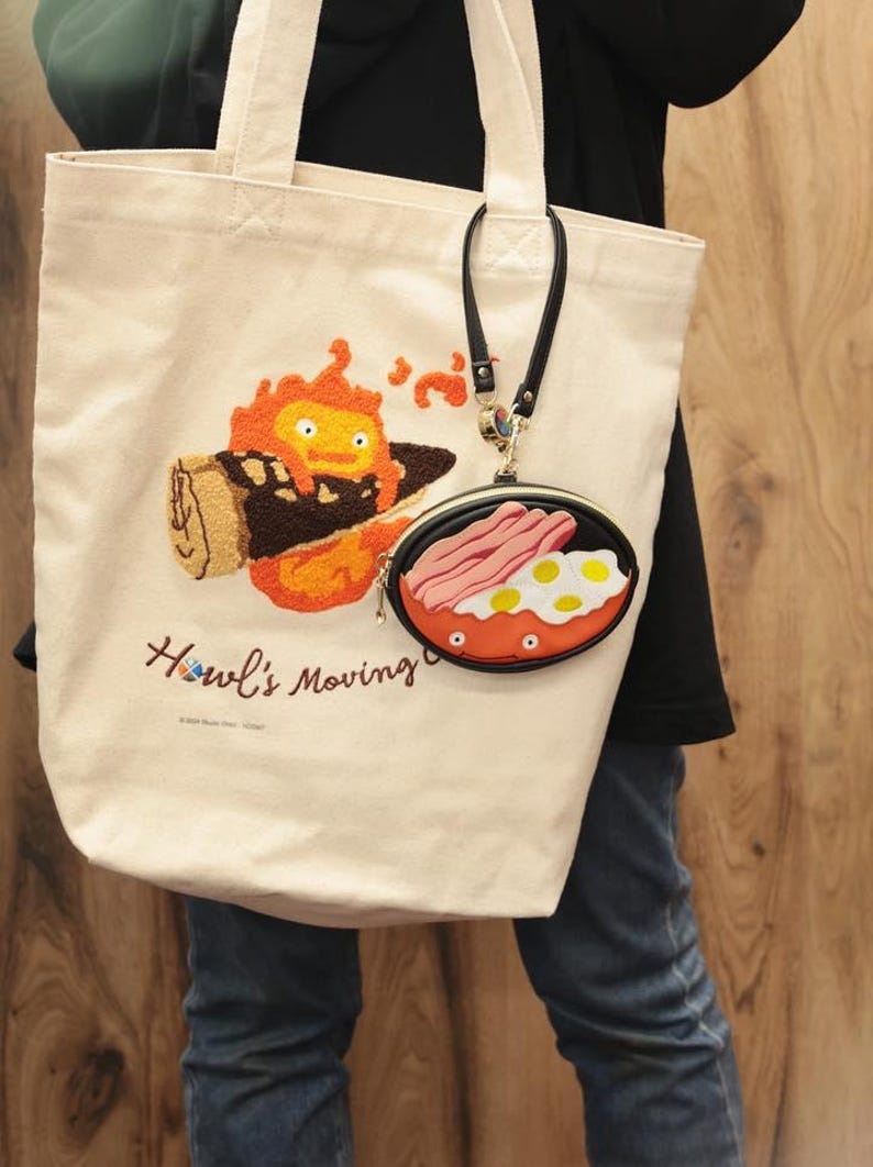 May include: Cream tote bag featuring an embroidered orange fiery character and the text "Howl's Moving". A small, round coin purse with a fried egg and bacon design is attached to the bag.