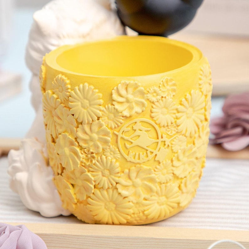 Puede incluir: Una maceta cil&iacute;ndrica de color amarillo brillante con una superficie texturizada cubierta de patrones florales en relieve. En la parte frontal presenta un emblema circular con la silueta de una figura sobre una escoba y el texto KIKI.