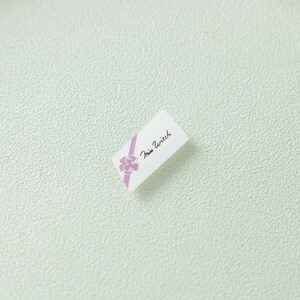 May include: A small rectangular white tag featuring a diagonal lavender ribbon graphic with a bow and the text Miss Witch printed in a black cursive script. The tag is oriented at an angle against a textured light green background surface.
