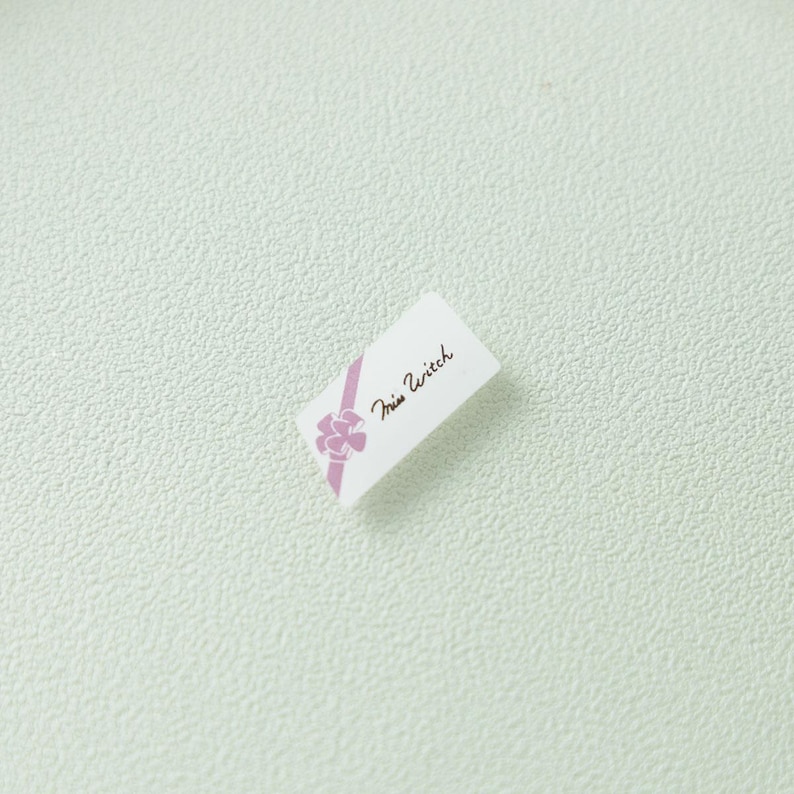 May include: A small rectangular white tag featuring a diagonal lavender ribbon graphic with a bow and the text Miss Witch printed in a black cursive script. The tag is oriented at an angle against a textured light green background surface.