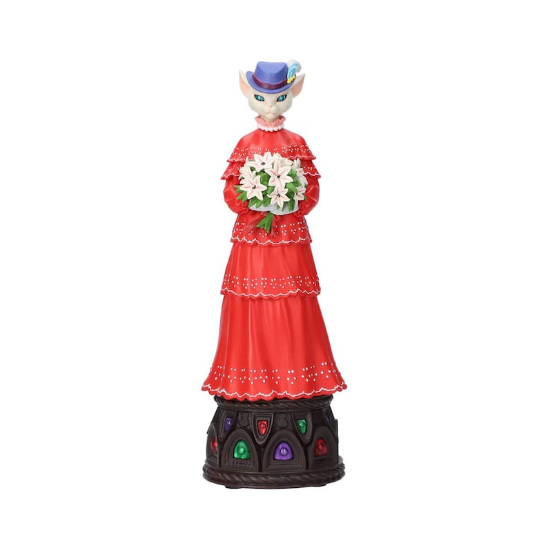 Puede incluir: Figura decorativa de un personaje felino con un vestido rojo y un sombrero azul, sosteniendo flores blancas. La figura tiene una base detallada con acentos coloridos.