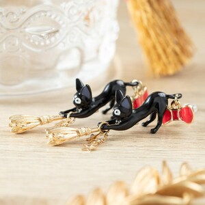 May include: A pair of black cat earrings with gold-colored broom charms and red bow accents. The earrings are set against a light wooden surface, with a blurred background featuring decorative elements.