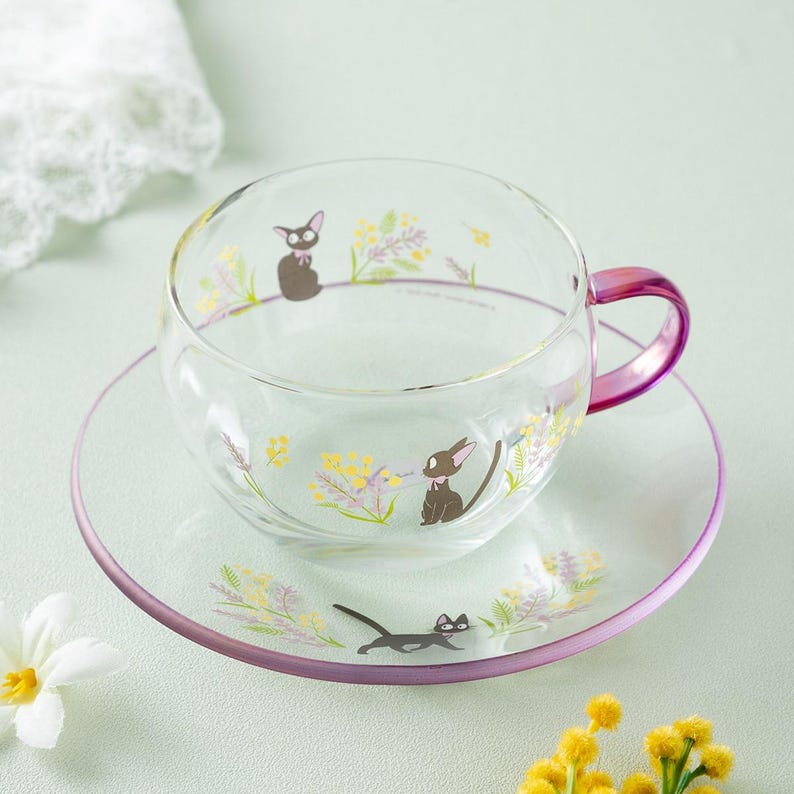 Op de afbeelding: Een theekop en schotel van helder glas met een paars handvat en rand. Het glas is versierd met illustraties van een kleine zwarte kat, gele mimosa-bloemen en lavendeltakjes, die zowel op de kop als op de schotel zijn afgebeeld.