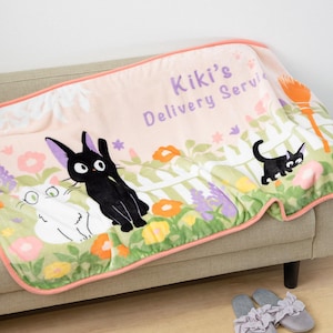 Puede incluir: Una manta de felpa con el texto "Kiki's Delivery Service". Muestra ilustraciones de gatos blancos y negros en un jardín floral, con un borde rosa y la inscripción.