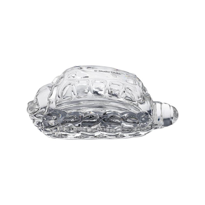 May include: A clear glass turtle figurine with textured shell details. The figurine has a rounded body and a small head and tail. The text "Studio Ghibli" is printed on the top of the figurine.