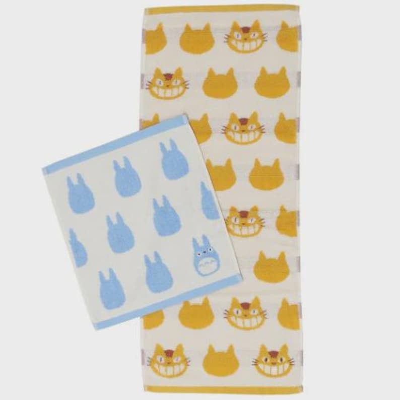 May include: Two patterned towels. One is rectangular with a repeating pattern of yellow cat faces and cat silhouettes on a cream background. The other is square with blue bunny-like characters on a cream background. Both have a cream border.
