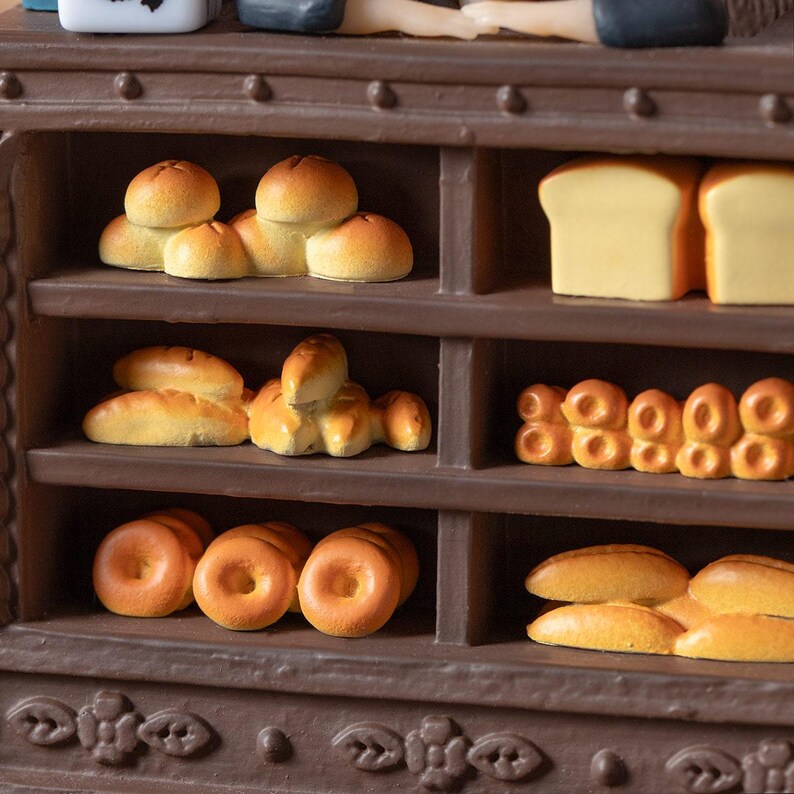 Pode incluir: Uma exibi&ccedil;&atilde;o de padaria em miniatura com v&aacute;rios produtos de panifica&ccedil;&atilde;o. As prateleiras est&atilde;o repletas de p&atilde;ezinhos redondos, p&atilde;es e bagels. Os itens s&atilde;o marrons dourados e dispostos em prateleiras marrons escuras, criando uma cena acolhedora.
