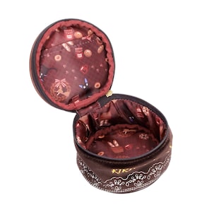 May include: A brown, round, open cosmetic bag with a zipper. The interior features a patterned fabric with bread, pastries, and a black cat. The exterior has white floral embroidery and the word "KIKI" in gold.