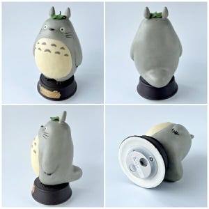 May include: A gray Totoro figurine with a cream-colored belly, black base, and a green leaf on its head. The figurine has a detailed design and is approximately 4 inches tall. The base has a gold-colored label.