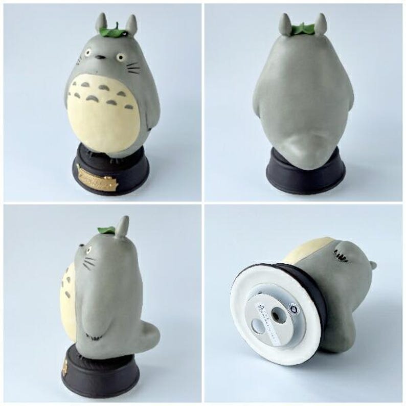May include: A gray Totoro figurine with a cream-colored belly, black base, and a green leaf on its head. The figurine has a detailed design and is approximately 4 inches tall. The base has a gold-colored label.
