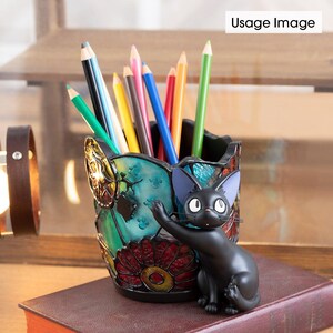 K&ouml;nnte beinhalten: Ein Bleistifthalter aus Buntglas mit einer schwarzen Katzenfigur. Der Halter hat ein florales Design mit roten, gelben und blauen Akzenten. Die Katze h&auml;lt eine Pfote hoch und hat ein schwarz-wei&szlig;es Gesicht.