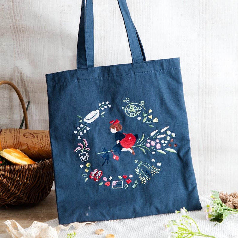 K&ouml;nnte beinhalten: Eine dunkelblaue Canvas-Tragetasche mit einem gestickten Design, das ein M&auml;dchen zeigt, das auf einem Besen mit einer Katze fliegt, umgeben von Blumen und anderen skurrilen Details. Der Text "KIKI" ist in einem Kreis gestickt.