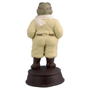 May include: A ceramic figurine depicting a figure in a vintage aviator outfit, seen from behind. The figure is dressed in a cream jumpsuit, brown boots, and a white scarf. It stands on a brown circular base.