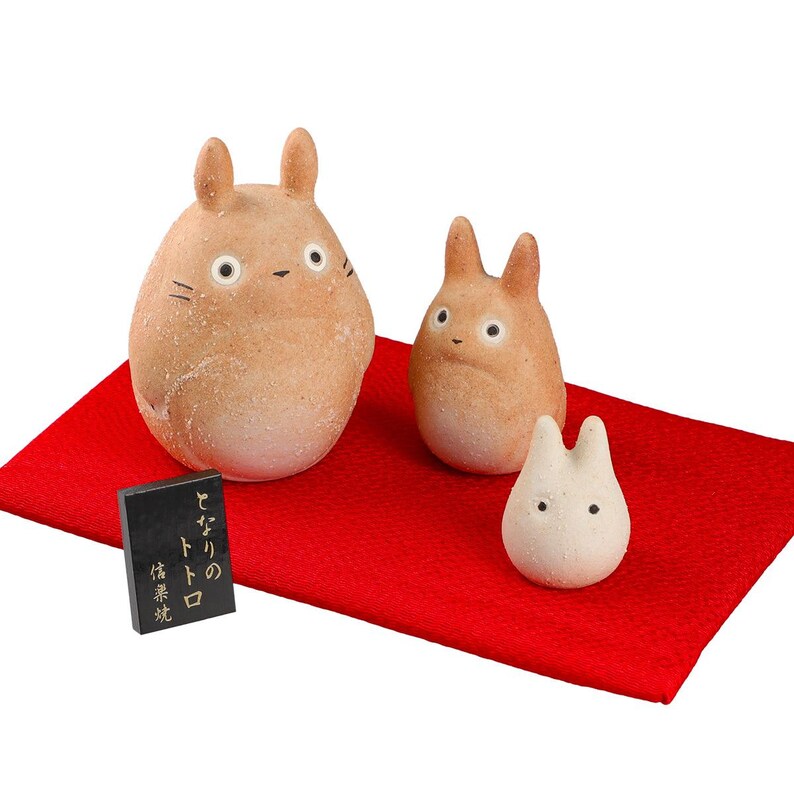 May include: Three ceramic Totoro figures in varying sizes, with tan bodies and black eyes, sit on a red cloth. A small black rectangular block with Japanese text is also present. The figures have a whimsical, cartoonish design.