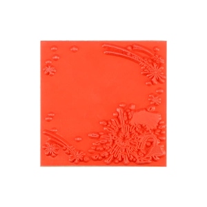 May include: A square, bright orange rubber stamp sheet featuring a raised, embossed design of floral elements and small circular dots arranged in the corners, creating a decorative pattern for crafting and stamping applications.