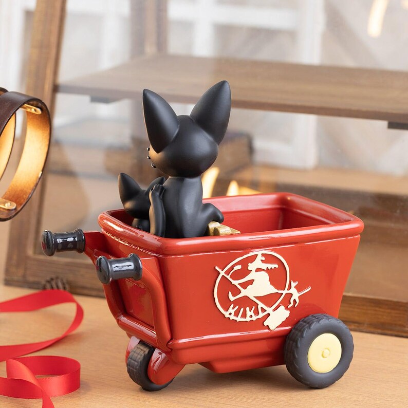 May include: A red decorative wagon with a black cat figurine inside. The wagon has a gold emblem of a witch on a broom. The wheels are black with yellow centers. The cat figurine is black.