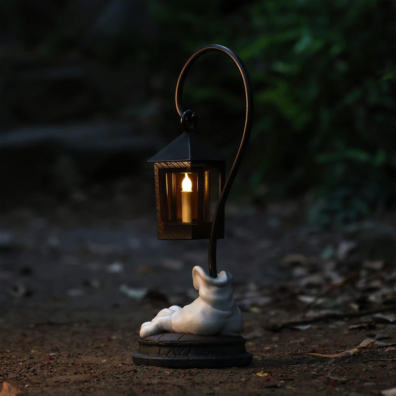 May include: A black metal lantern with a curved support, featuring a small, lit candle inside. The base is a white sculpture of a reclining figure. The lantern is designed for outdoor use and has a vintage aesthetic.