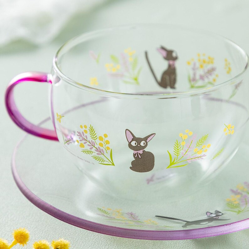 Op de afbeelding: Een set van een doorzichtige glazen theekop en schotel met een felroze handvat en rand. Het glas is versierd met illustraties van kleine zwarte katten met roze strikjes, omringd door trossen gele en paarse bloemen en groen blad.