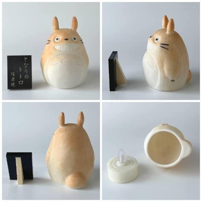 Może przedstawiać: Beżowo-biała ceramiczna figurka Totoro ze spiczastymi uszami, szerokim uśmiechniętymi ustami i wąsami. Zestaw zawiera mały czarny prostokątny znak z japońskim tekstem oraz białą zasilaną bateryjnie świeczkę LED, kt&oacute;ra mieści się wewnątrz.