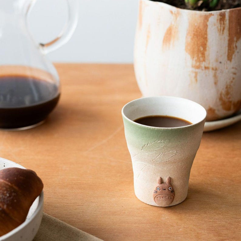May include: A ceramic coffee cup with a green and white gradient, filled with dark coffee. The cup features a Totoro figure on the base and leaf-like etchings. A glass carafe and a croissant are also visible.