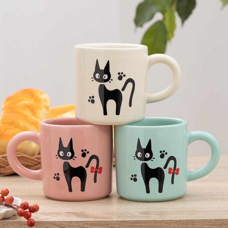 May include: Three ceramic mugs in cream, pastel pink, and light blue, each featuring a black cat illustration with a small red bow on the pink and blue mugs, and paw prints surrounding the cat figures on all three mugs with rounded handles.
