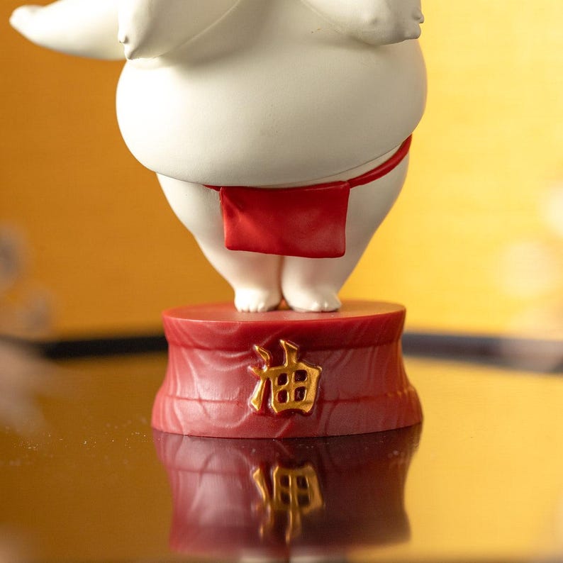 Peut inclure: Figurine blanche repr&eacute;sentant un lutteur de sumo avec un pagne rouge. La figurine est pos&eacute;e sur un socle rouge avec des caract&egrave;res japonais dor&eacute;s. Le fond est un d&eacute;grad&eacute; de jaune et d'or.