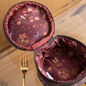 May include: Open, round, brown zippered pouch with a patterned interior featuring bread, jam, and cat illustrations. A gold fork is in the foreground. The pouch is open, revealing the interior design.