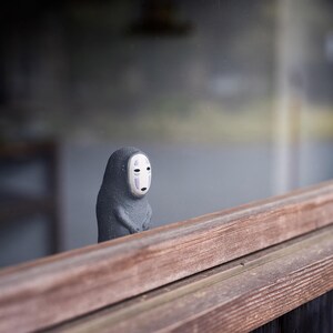 Puede incluir: Una peque&ntilde;a figura de color gris oscuro con textura y un rostro liso de color blanco p&aacute;lido similar a una m&aacute;scara, con dos l&iacute;neas verticales moradas bajo los ojos y una peque&ntilde;a boca negra. La figura se sit&uacute;a tras un borde de madera horizontal.