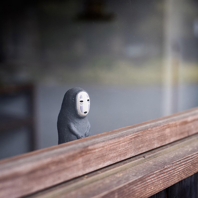 Puede incluir: Una peque&ntilde;a figura de color gris oscuro con textura y un rostro liso de color blanco p&aacute;lido similar a una m&aacute;scara, con dos l&iacute;neas verticales moradas bajo los ojos y una peque&ntilde;a boca negra. La figura se sit&uacute;a tras un borde de madera horizontal.