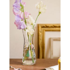 Puede incluir: Un jarr&oacute;n de vidrio transparente con un dise&ntilde;o curvo, que contiene un ramo de flores de guisante de olor de colores pastel. El jarr&oacute;n tiene una ilustraci&oacute;n decorativa y texto. Las flores incluyen tonos blancos, rosas y morados.