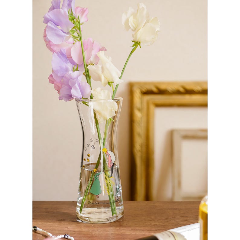Puede incluir: Un jarr&oacute;n de vidrio transparente con un dise&ntilde;o curvo, que contiene un ramo de flores de guisante de olor de colores pastel. El jarr&oacute;n tiene una ilustraci&oacute;n decorativa y texto. Las flores incluyen tonos blancos, rosas y morados.