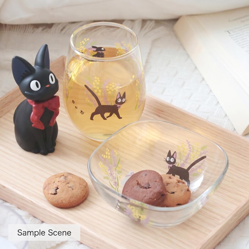 May include: A clear glass tumbler and a square glass bowl, both featuring a black cat illustration with yellow and purple floral accents. A black cat figurine with a red bow sits beside the glass, and two round chocolate chip cookies are present.