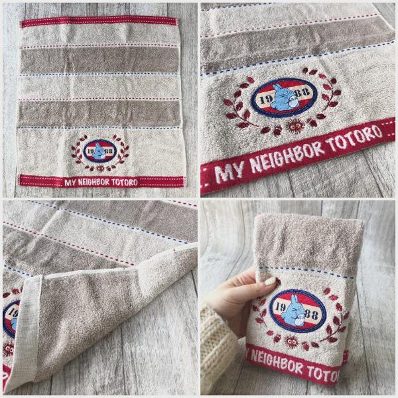 Puede incluir: Toalla a rayas beige y marr&oacute;n con un dise&ntilde;o bordado. El dise&ntilde;o incluye un personaje Totoro azul, el a&ntilde;o "1988" y el texto "MY NEIGHBOR TOTORO" en rojo.