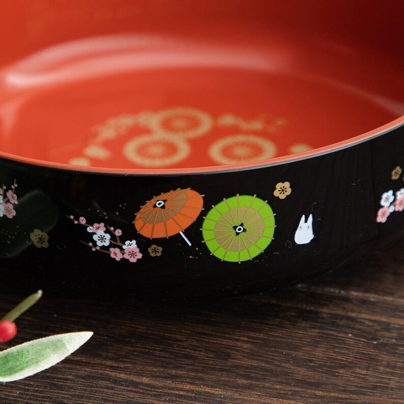 May include: A black and orange decorative bowl with a detailed design. The bowl features a vibrant orange interior and a black exterior adorned with colorful illustrations of umbrellas, flowers, and a white cartoon character. The bowl is likely used for serving food or as a decorative piece.