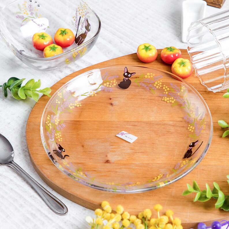 May include: A clear glass plate and a matching glass bowl featuring a printed design of a black cat with a pink bow, surrounded by yellow and purple floral sprigs. The plate has a small rectangular white label in the center with black text.
