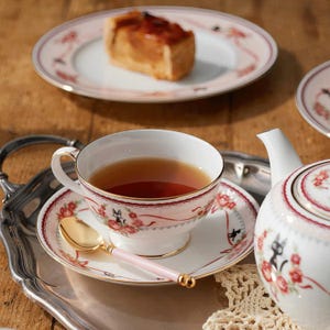 May include: A tea set with a cup, saucer, and teapot, featuring a floral and ribbon design with a black cat motif. A gold spoon with a pink handle rests on the saucer. A plate with a slice of cake is in the background.