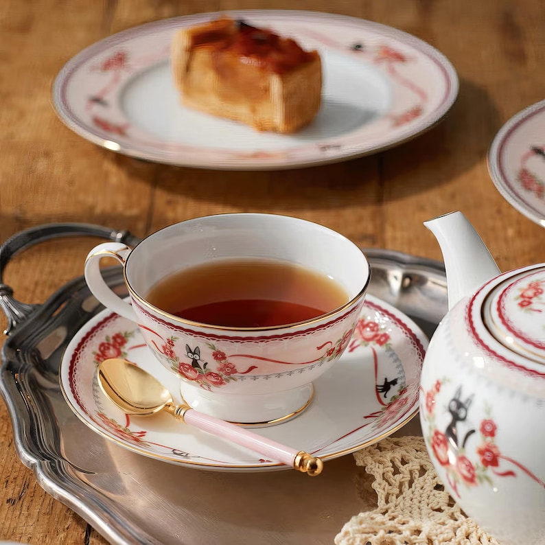 May include: A tea set with a cup, saucer, and teapot, featuring a floral and ribbon design with a black cat motif. A gold spoon with a pink handle rests on the saucer. A plate with a slice of cake is in the background.