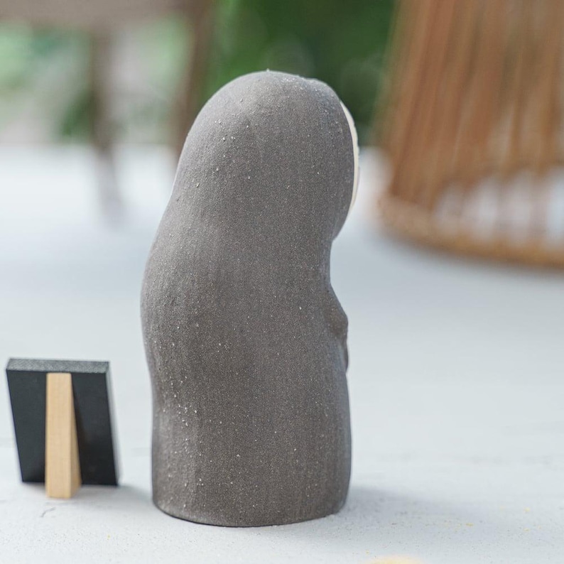 Puede incluir: Una peque&ntilde;a figura de color gris oscuro con textura, forma redondeada con capucha y un rostro blanco liso. Junto a la figura hay un letrero rectangular negro en miniatura sostenido por un caballete de madera clara, sobre una superficie plana.