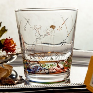 May include: A clear glass tumbler with a gold-colored illustration of a character from an animated film. The design features a character holding a small creature, with shooting stars and a decorative band at the base. The glass is on a silver tray.